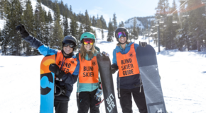 Three blind snowboarders in vests waving to camera on the slopes