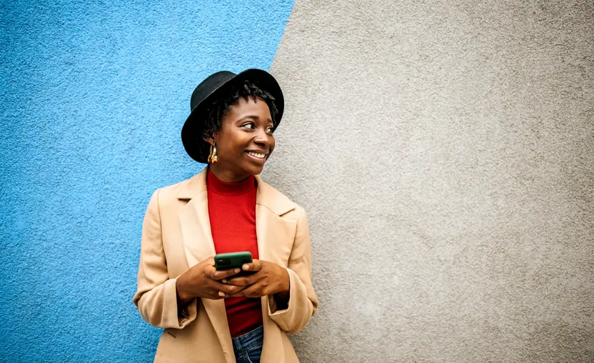 This image depicts a young Black woman in casual clothes leaning against a multicolored wall with a phone in her hands.