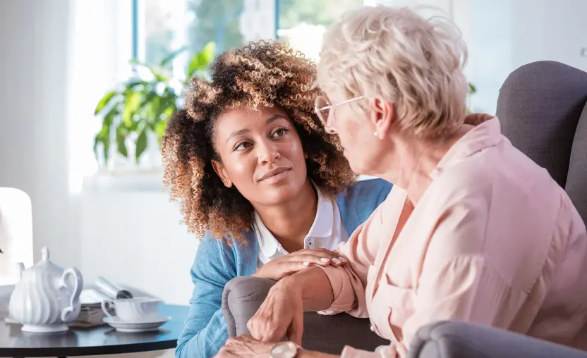A human caregiver talks with a senior woman sitting in a living room chair, after connecting through case management software for human services.