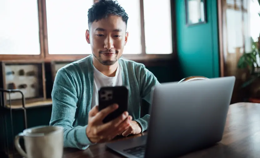 A confident young Asian man looking at his smartphone while working on a laptop computer in his home office, exploring ways to turn Facebook likes into donations.