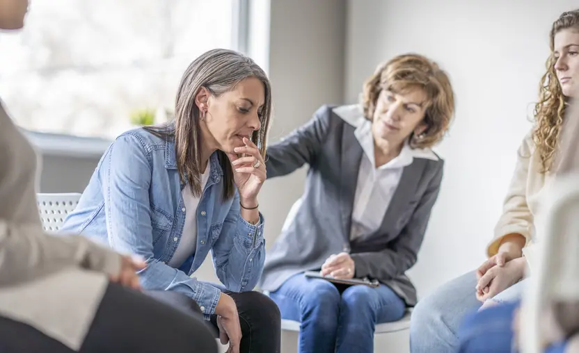 A female therapist addresses barriers to mental health and reaches her hand out to comfort a woman sitting next to her during a group therapy session.