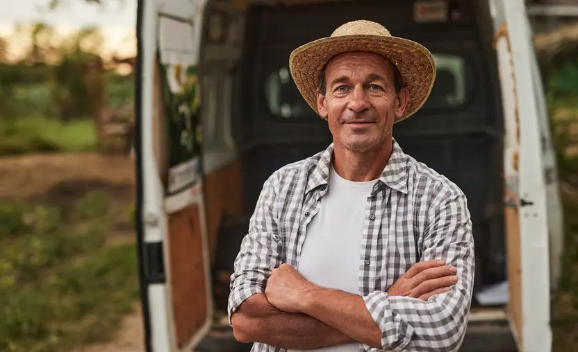 A smiling farmer stands with crossed arms against blurred van prepared for delivery of products. 