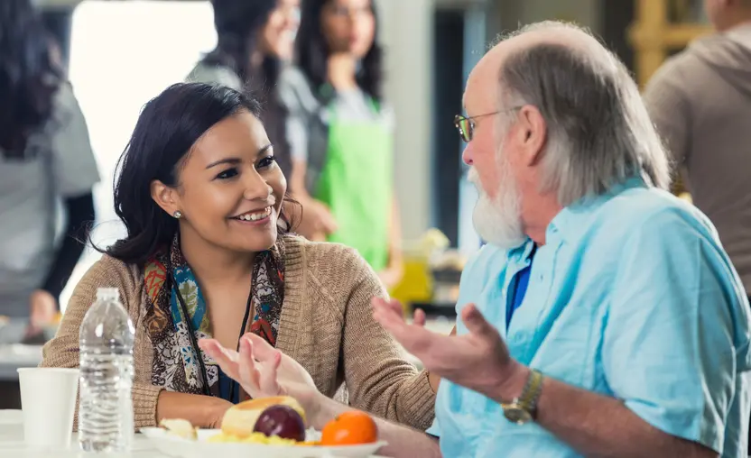 Young adult Hispanic woman is sitting at table in soup kitchen and talking with senior Caucasian man. Man has grey hair and beard. They are discussing something while senior enjoys healthy meal. Volunteers are serving food in backgound.
