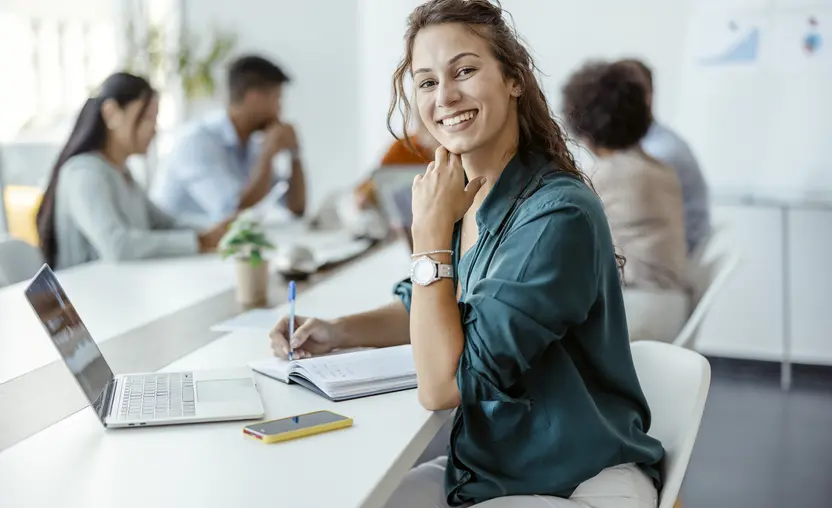 A female employee smiling in the office with her colleagues in the background. 
