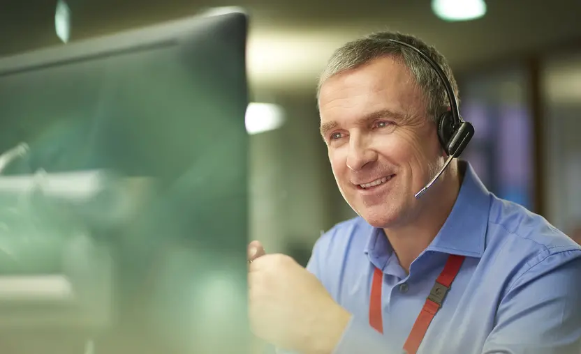 A call center phone representative with short gray hair chats on the phone at his desk. He is discussing something on the phone in a friendly way. Behind him is an out of focus office interior.
