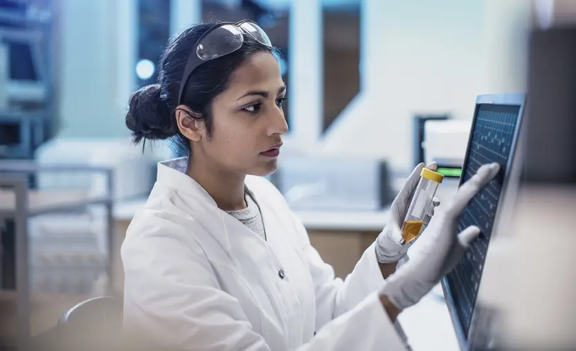 A scientist works in a lab using a touch screen computer. She has long dark hair and is wearing a white coat.