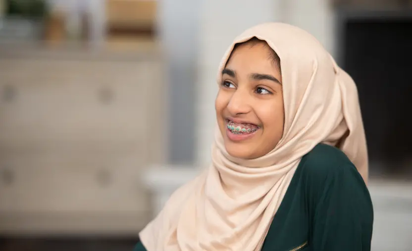 A young teenage Muslim girl sits in the comfort of her home while posing for a portrait. She is dressed casually and wearing a Hijab. She is seated to the right and sitting sideline as she looks to the left and smiles.