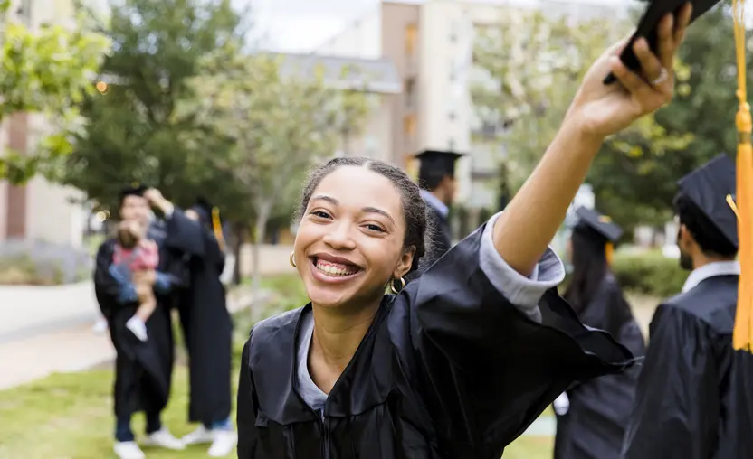 The joyful young adult graduate raises her cap in her hand as she poses for a photo.