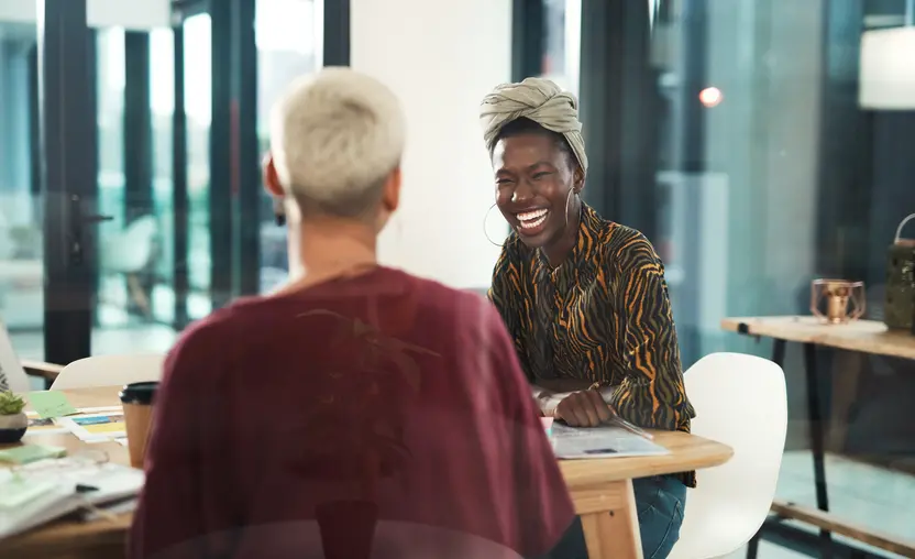 Black woman with head wrap laughing while talking to colleague at table in professional environment