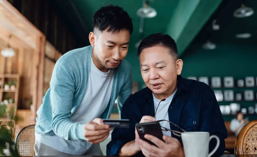 A father and his adult son, both with short dark hair and in blue shirts, read together from the father’s smartphone. They are indoors at a table in a dark green office.