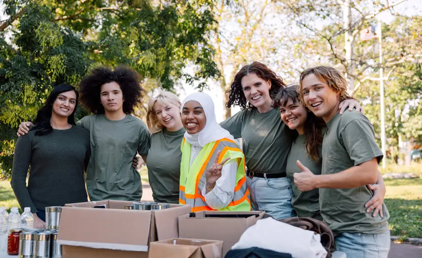 A group of seven nonprofit volunteers gather behind a table of canned food and smile at the camera for a photo. All but one are wearing green long- or short-sleeved t-shirts. One is wearing a green high-visibility reflective vest and a long-sleeved white shirt.