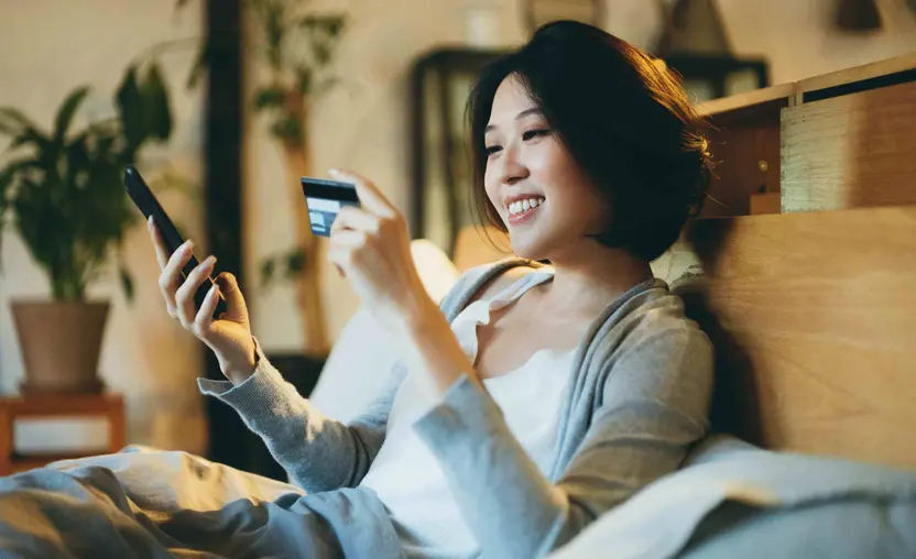 A smiling young person with dark hair is holding their credit card in one hand and their smartphone in the other. They are wearing a white shirt and a gray cardigan.