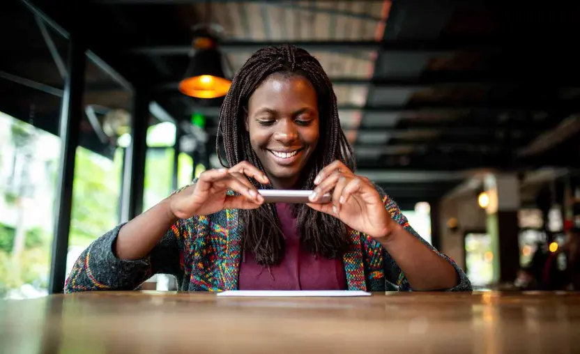A young Black woman in a colorful sweater is seated at a wooden bar and smiling as she uses her smartphone to take a photo of a piece of paper. She might be using a QR code.