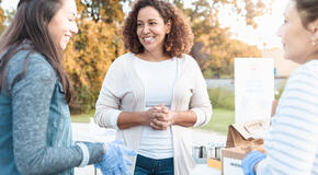 Three women smile and chat while volunteering at a community food drive, representing the importance of starting an employee volunteer program.