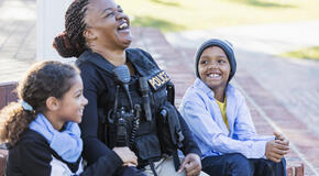A female police officer is conversing with a boy and his sister, sitting side by side on steps outside a building, laughing.