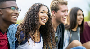 A girl of African American descent smiles with laughter while sitting in a row of teens with the arms around each other's shoulders. They are laughing too.