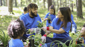 A group of environmental volunteers in matching bright blue t-shirts plant flowers, trees, and plants at a local park during the spring.