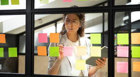 A businesswoman stands behind a glass wall and conducts a brainstorming session by placing multicolored sticky notes with ideas on the wall. 