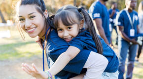 Young girl rides piggyback on her big sister's back during a community cleanup event. They are smiling at the camera.