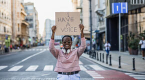 A passionate nonprofit advocate stands at a crosswalk, smiling and holding up a sign that says "I have a voice."