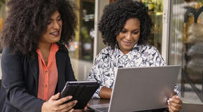 Two women work together at a cafe on a digital tablet and a laptop.