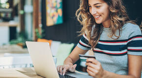 This photo of a smiling young woman holding a credit card and typing on a laptop represents the concept of a sustainer upsell lightbox.