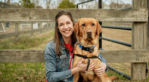 A brown-haired woman in a red t-shirt, black pants, and a denim jacket sits on the grass in front of a wood fence hugging a medium sized reddish-brown dog. She is smiling at the camera.