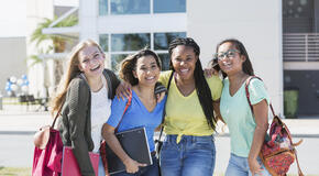 A group of f teenage girls, standing in front of a school building carrying book bags and notebooks.