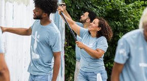 A diverse group of cheerful volunteers roll paint over a graffiti covered wall.