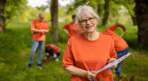 A volunteer stands in a forest with a clipboard while four other volunteers work to plant trees, demonstrating the value of volunteer time.