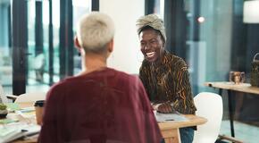 Black woman with head wrap laughing while talking to colleague at table in professional environment