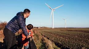 A father and son are leaning against a fence in a field together, looking at the wind turbines.