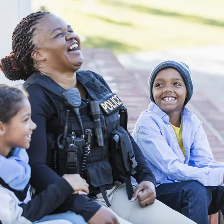 A female police officer is conversing with a boy and his sister, sitting side by side on steps outside a building, laughing.