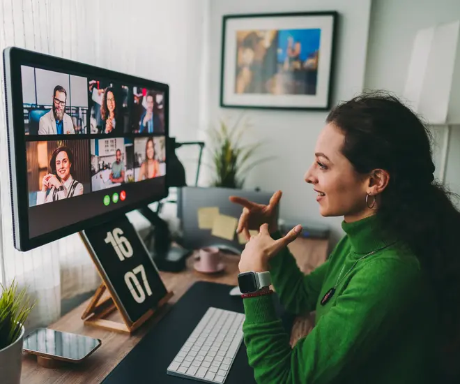 A person with long brown hair in a green turtleneck and a smartwatch discusses their work on a virtual meeting with six coworkers.