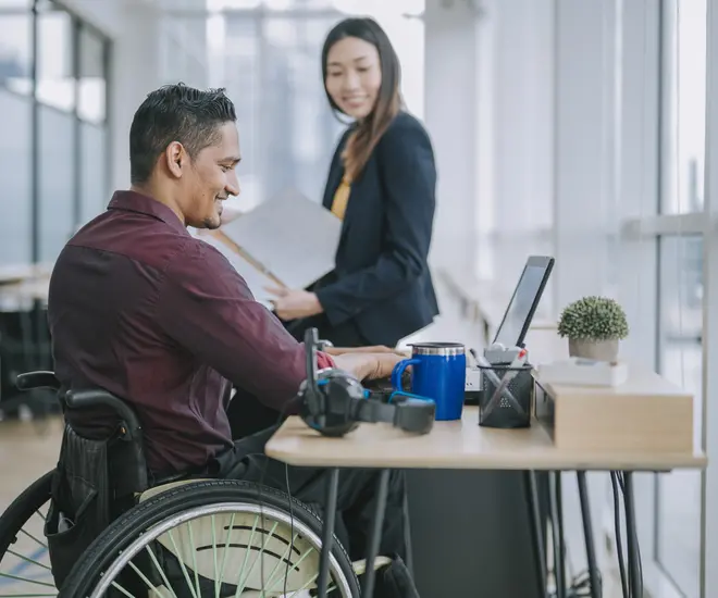 Man in wheelchair working on a computer while talking with a female colleague.