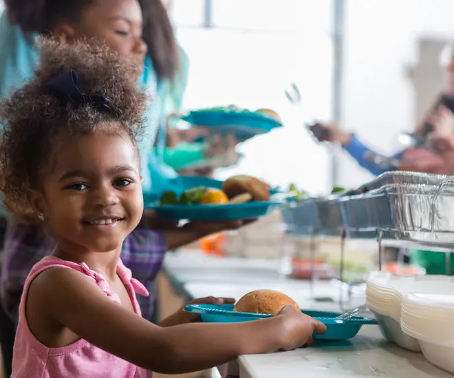 Cheerful African American little girls smiles while in line in a soup kitchen. She is holding a plate full of healthy food. Her family is in line behind her.