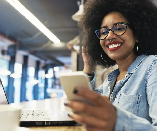 A Black woman with dark brown hair, a septum piercing, red lipstick, and a blue denim shirt smiles at her smart phone in an office.