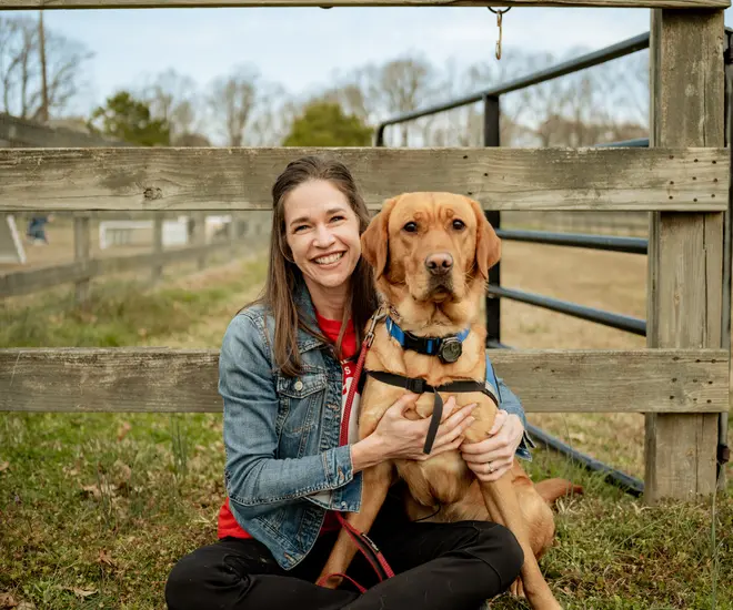 A brown-haired woman in a red t-shirt, black pants, and a denim jacket sits on the grass in front of a wood fence hugging a medium sized reddish-brown dog. She is smiling at the camera.