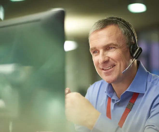 A call center phone representative with short gray hair chats on the phone at his desk. He is discussing something on the phone in a friendly way. Behind him is an out of focus office interior.