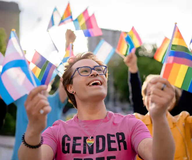 A person with short hair in a pink shirt is smiling and waving a rainbow flag and a trans pride flag, which is pink, blue, and white.