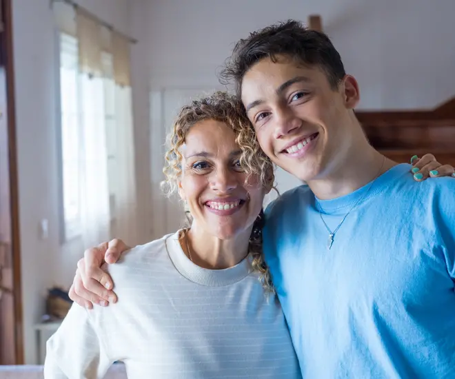 A mother and her teenage son smile at the camera. She is wearing a white shirt and has blonde curly hair, and he has wavy brown hair and is wearing a blue t-shirt.