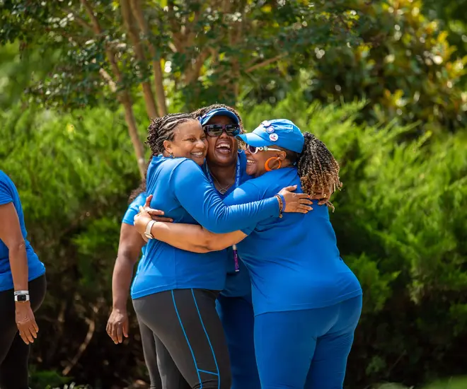 Three Black women wearing their royal blue GirlTrek member shirts embrace and laugh together. They are out on a GirlTrek walk together, it’s a sunny day, and there are trees in the background.