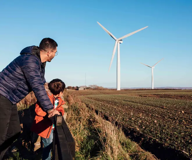 A father and son are leaning against a fence in a field together, looking at the wind turbines.