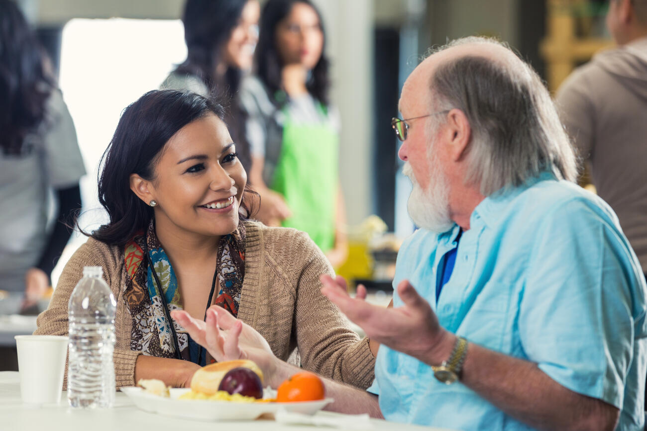Young adult Hispanic woman is sitting at table in soup kitchen and talking with senior Caucasian man. Man has grey hair and beard. They are discussing something while senior enjoys healthy meal. Volunteers are serving food in backgound.