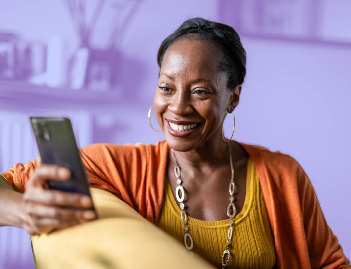 Smiling woman in yellow top and orange cardigan looks at her smartphone. Blurred purple background with yellow couch.