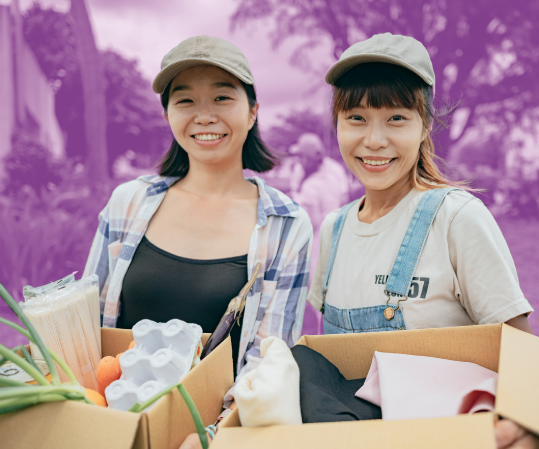 Two smiling volunteers holding boxes filled with food and supplies during an outdoor community event.