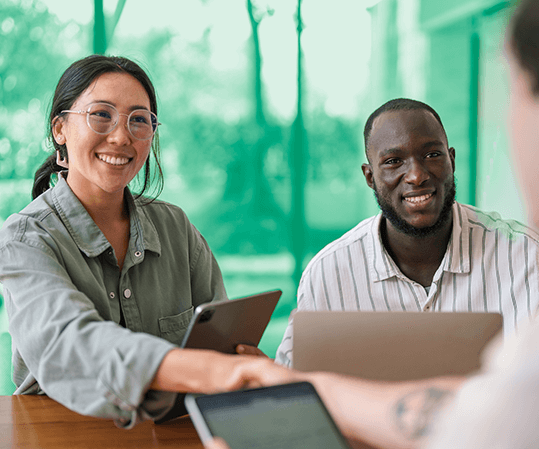Two smiling professionals seated at a table with laptops and tablets, shaking hands with a third person across from them.