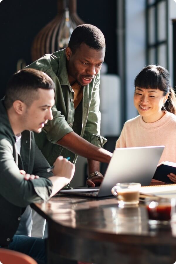A small group of professionals collaborate around a table, reviewing information together on a laptop in a bright workspace.