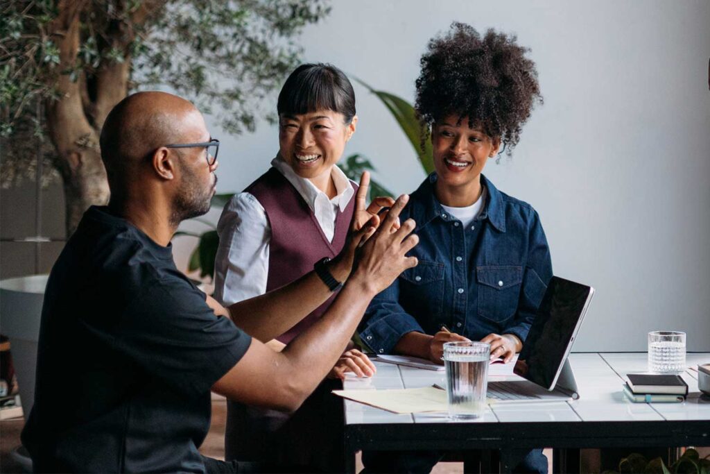 Three professionals collaborate at a table, discussing ideas while reviewing notes and a laptop in a bright workspace.