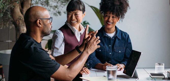 Three professionals collaborate at a table, discussing ideas while reviewing notes and a laptop in a bright workspace.